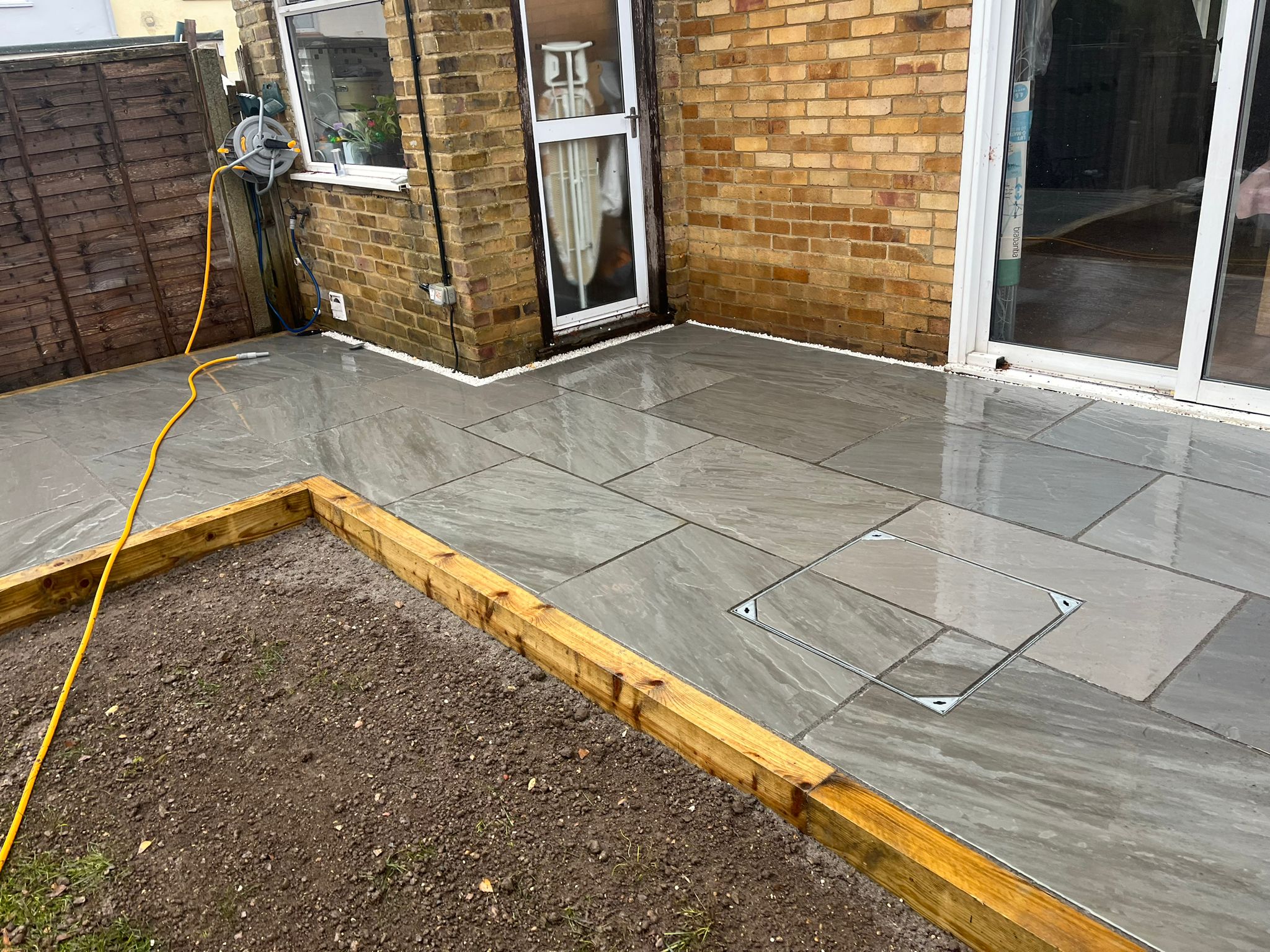 An image showing a newly laid patio in a back garden. Grey shiny slabs with a recessed drain.